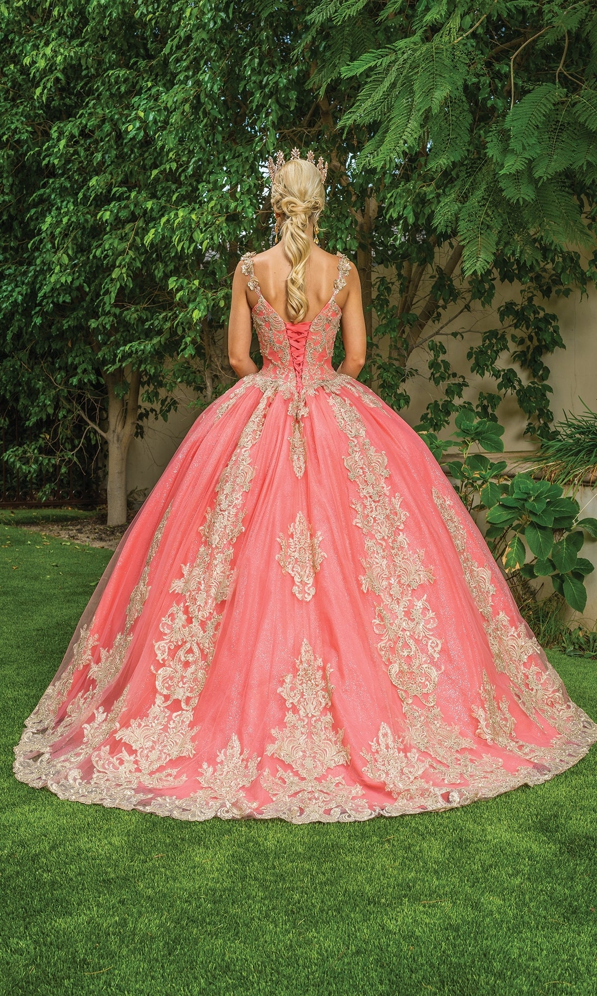 A woman stands on grass with her back to the camera, wearing the Coral Quinceanera Dress 1629—a coral pink ball gown with a full skirt and gold lace embroidery—plus a crown, surrounded by trees and greenery.